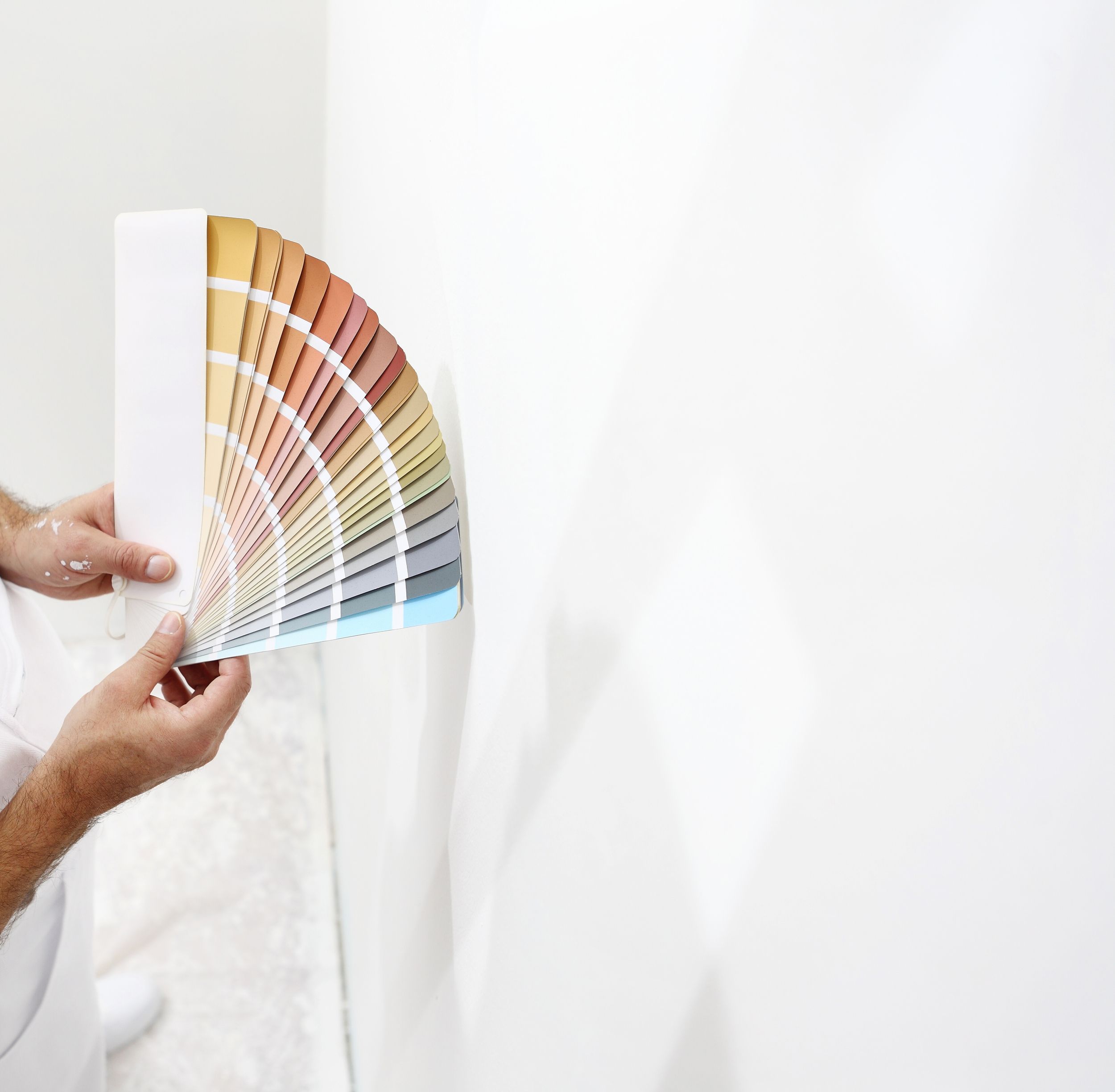 Eine Person hält ein Farbmuster-Fächer mit verschiedenen Farbtönen von Gold, Beige, Rosa bis Blau gegen eine weiße Wand.