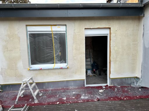 Hausfassade mit unvollendeter Putzarbeit, Leiter und offener Tür im Vordergrund.
